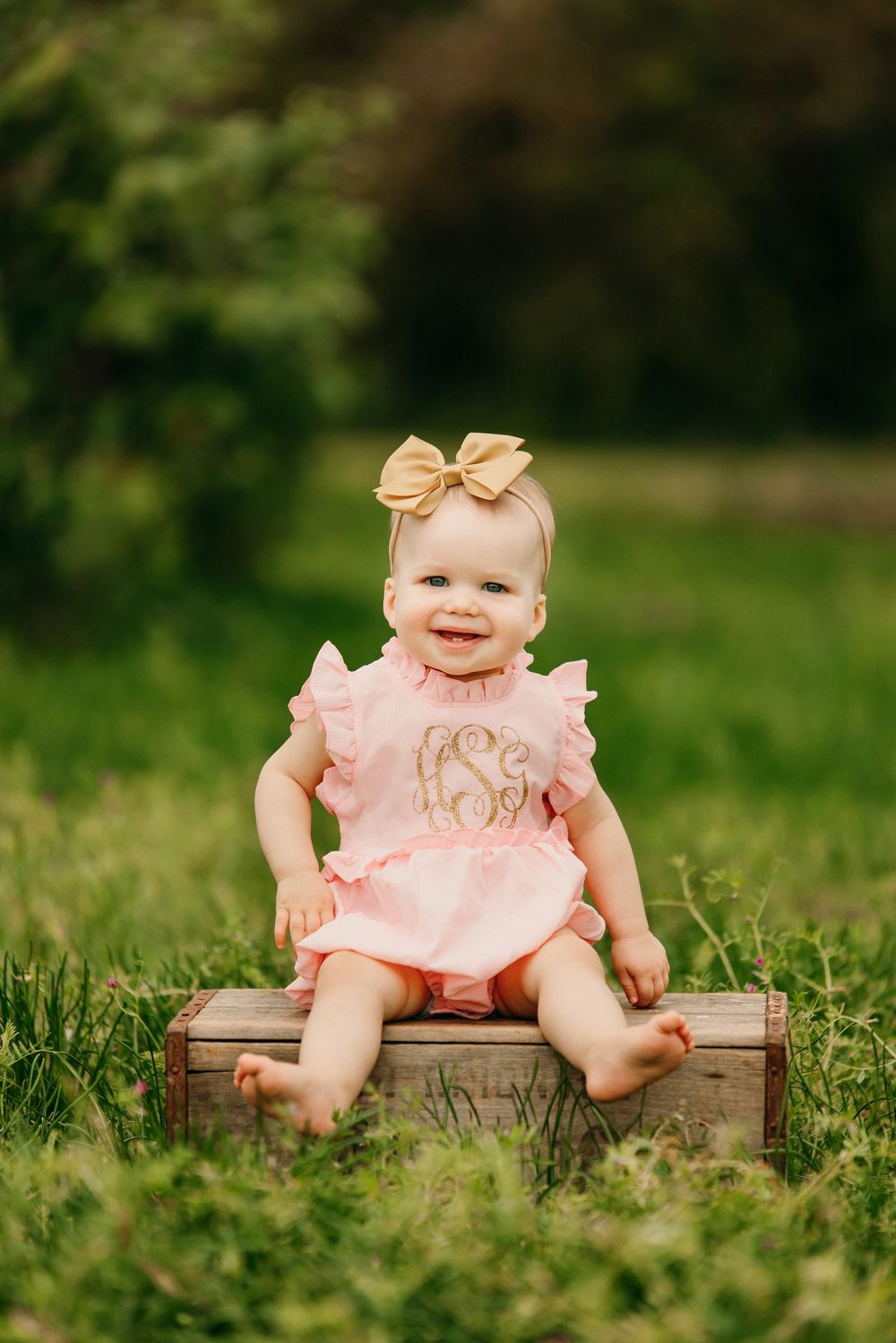 Baby wearing peach dress and bow sitting on wooden platform in grass.