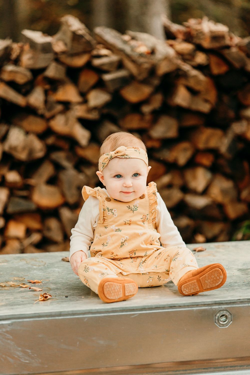 Baby in peach-colored outfit sits on wooden bench with stacked firewood in background.