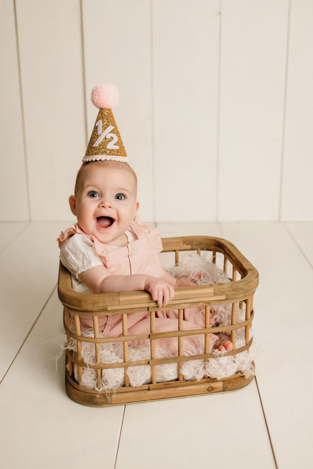 Baby wearing pink outfit sits in wooden basket against white backdrop.