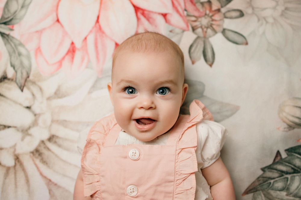 Baby in pink dress poses against floral backdrop.