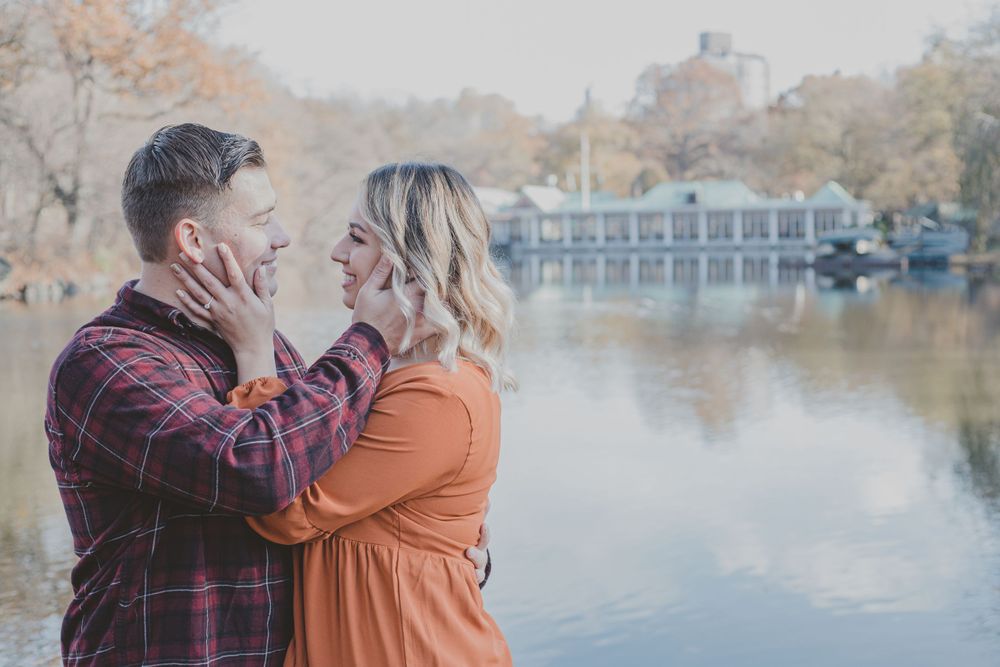A couple poses for engagement photos by a lake with fall colors in the background.