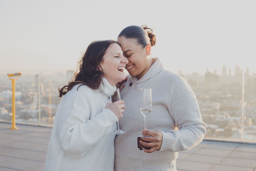 A couple sharing a joyful moment in white sweaters with city skyline in background.