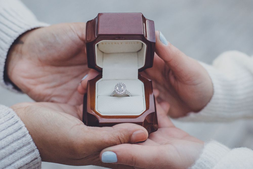 An engagement ring in burgundy velvet box held by hands wearing cozy sweaters.