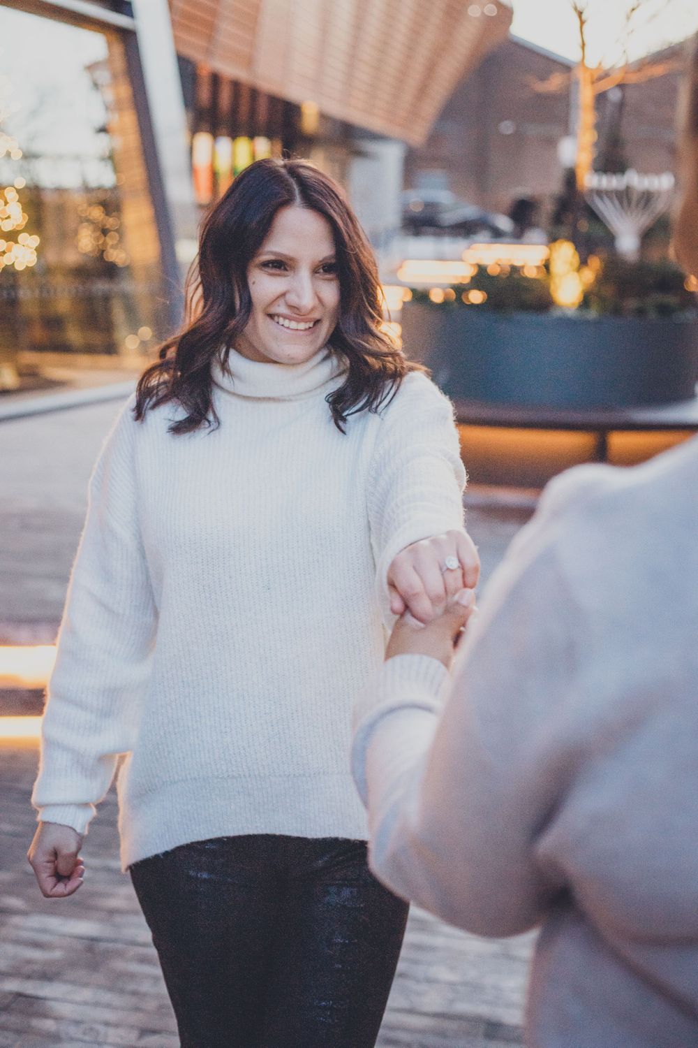 A person in white sweater smiling while showing off engagement ring outdoors.