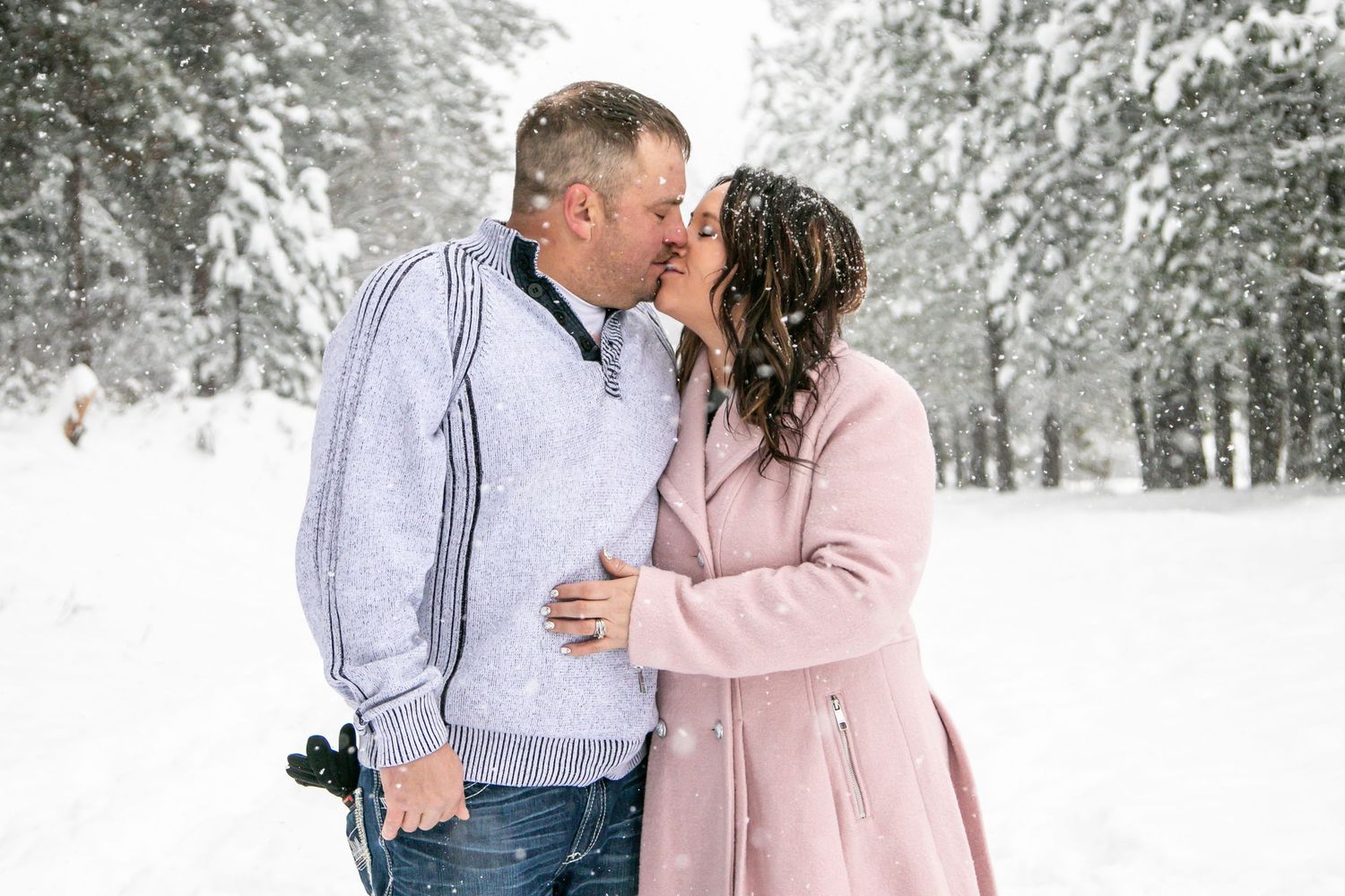 A couple shares an intimate moment in a snowy forest setting wearing light colored winter clothing.