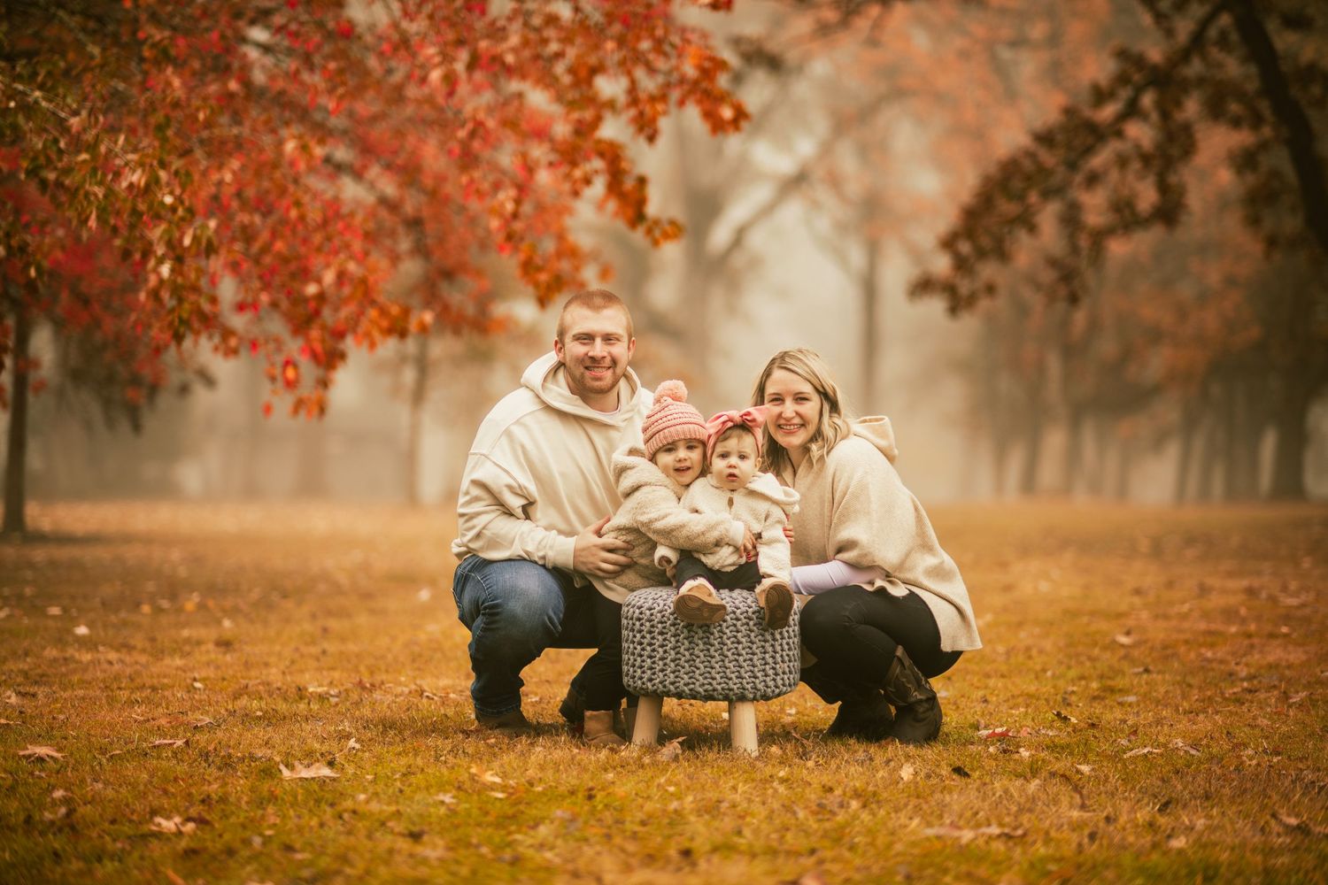 mom and dad crouching down next to daughter sitting on stool. other daughter hugging her sister. it is fall.