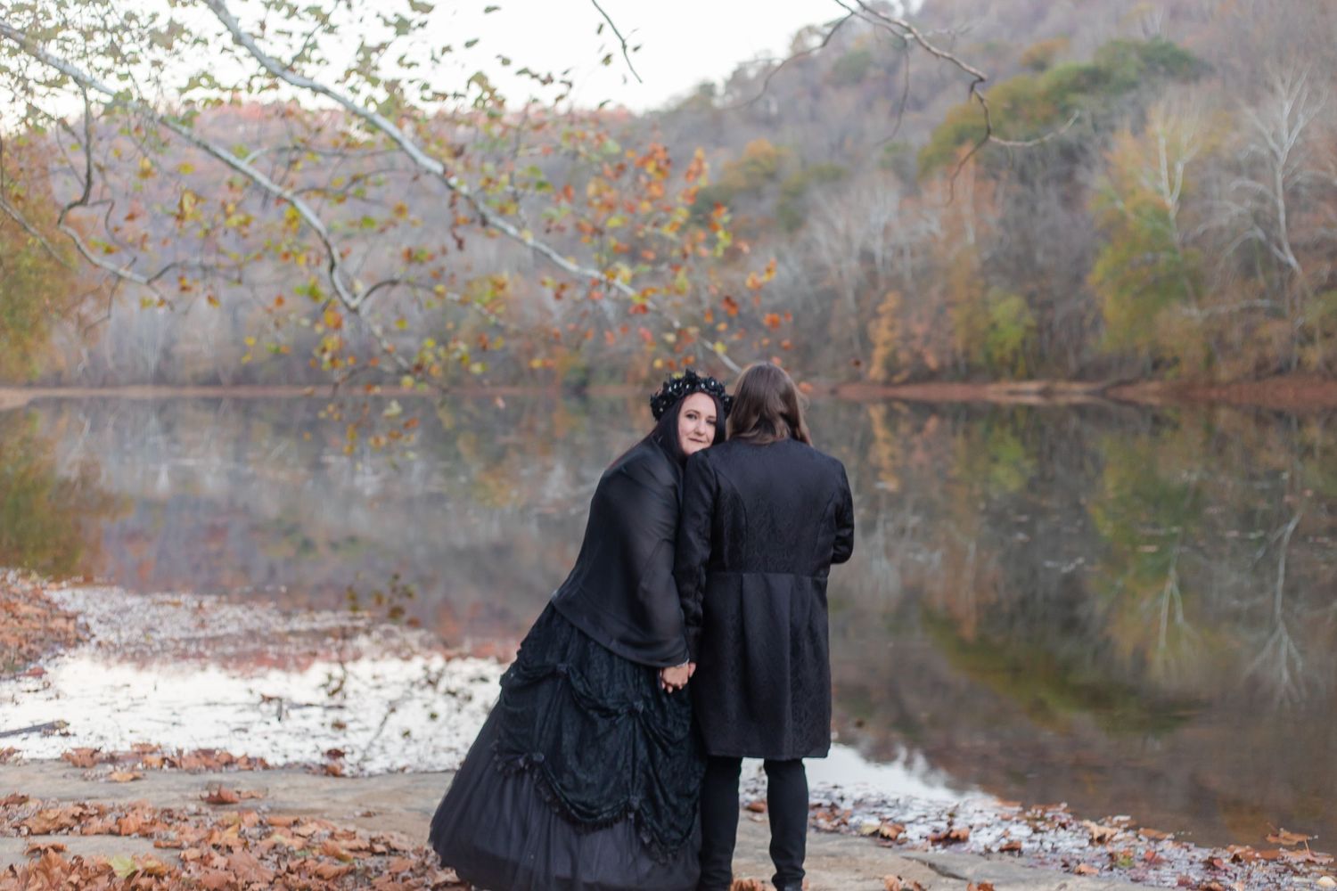 People in dark Victorian-inspired clothing gather by a lakeside on a fall day.