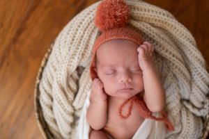 Sleeping newborn wearing knit hat with pom pom nestled in cream blanket.