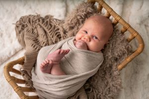 Newborn baby wrapped in beige fabric sleeps peacefully in wooden chair with fuzzy blanket.