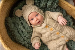 Baby dressed in beige knit sweater lying in woven basket against textured backdrop.