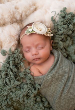 Peaceful newborn wrapped in gray swaddle with floral headband among dusty green foliage.