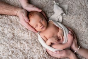 Newborn baby swaddled in white fabric lying on plush carpet with gentle hands supporting.