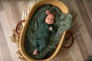 Newborn baby wearing green sleeper rests in round wicker basket with cozy textured blanket.
