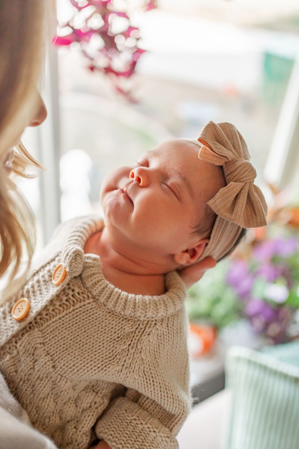 Sleeping newborn in cream knit sweater wearing delicate bow headband near vibrant flowers.