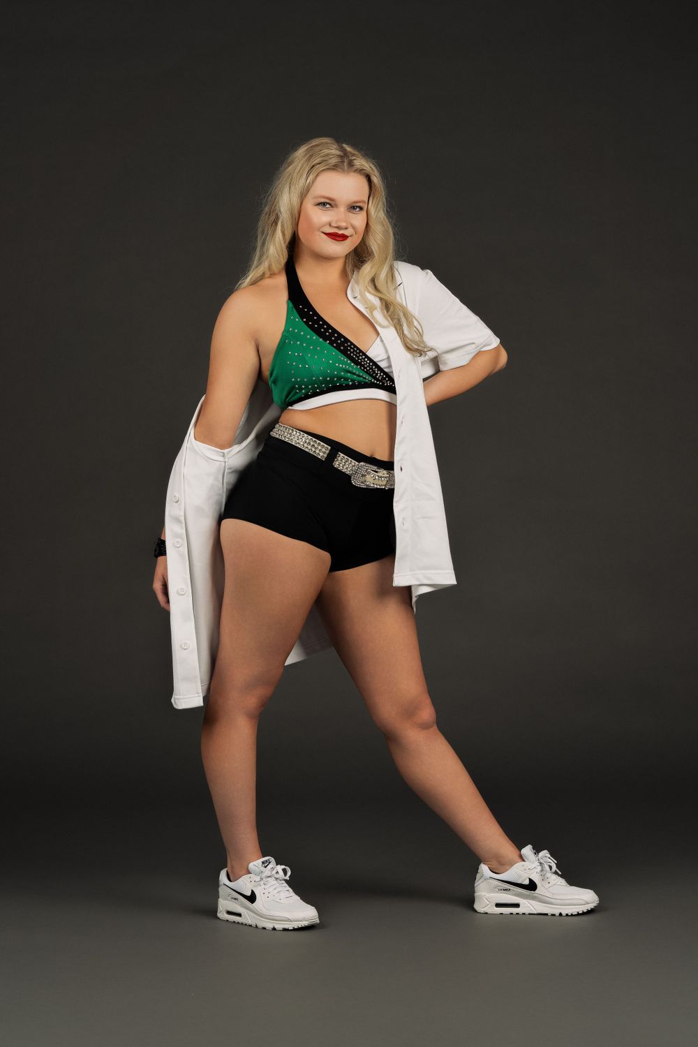 College dancer in green crop top, black shorts and white sneakers strikes dynamic pose against dark background.