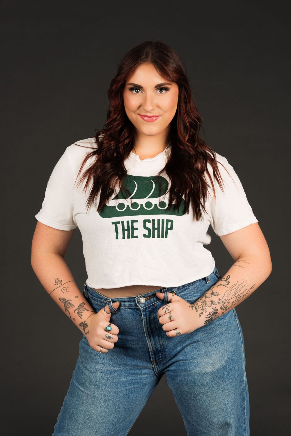 Dancer in a white crop top with The Ship logo and blue jeans poses confidently against dark background showing arm tattoos.