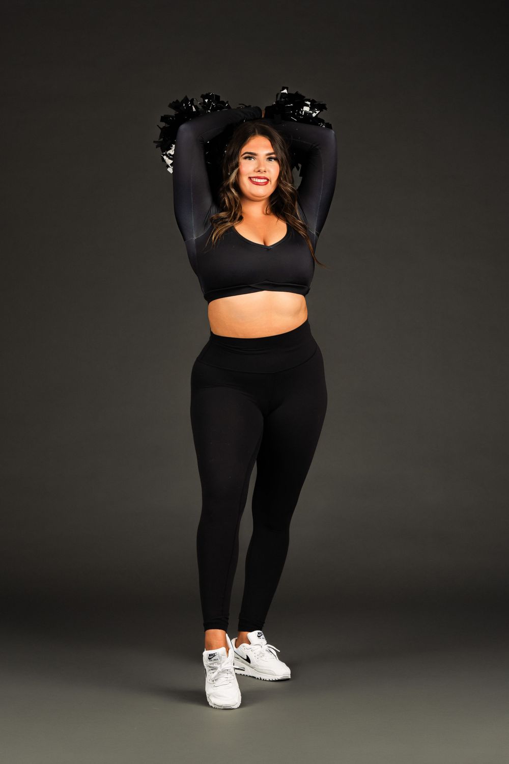 College Dancer in all-black workout attire with long sleeves and white sneakers strikes a pose against dark backdrop.