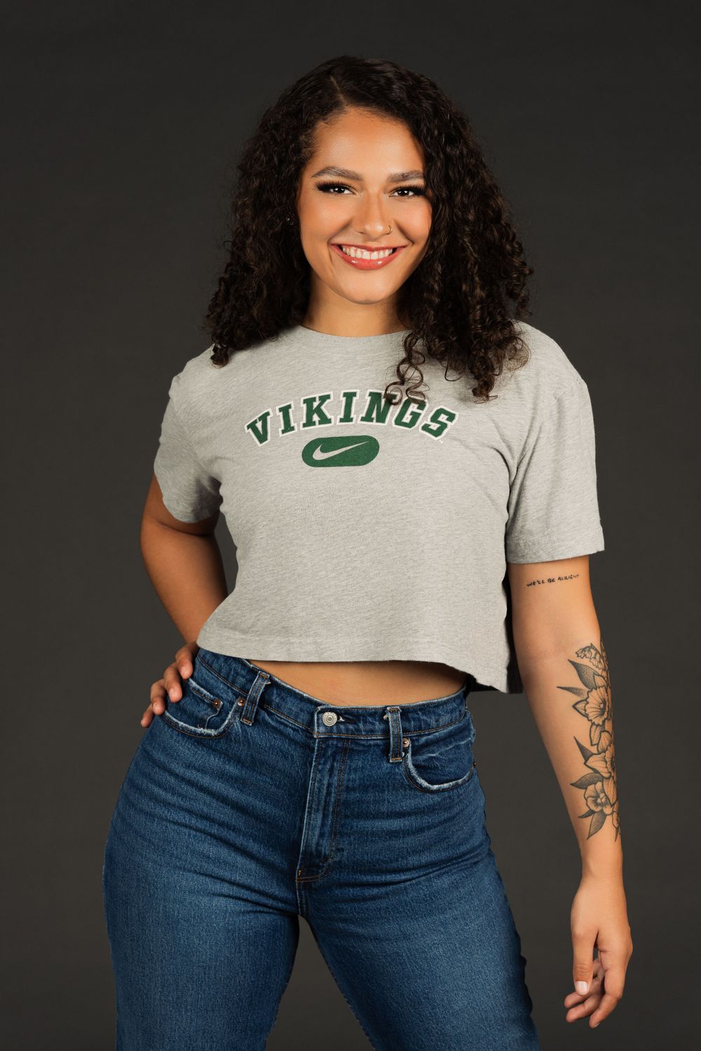 PSU college dancer in a gray cropped Vikings t-shirt and blue jeans stands against dark background with arm tattoo visible.
