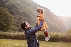 Playful moment captured as parent lifts child wearing orange dress in sunlit meadow.