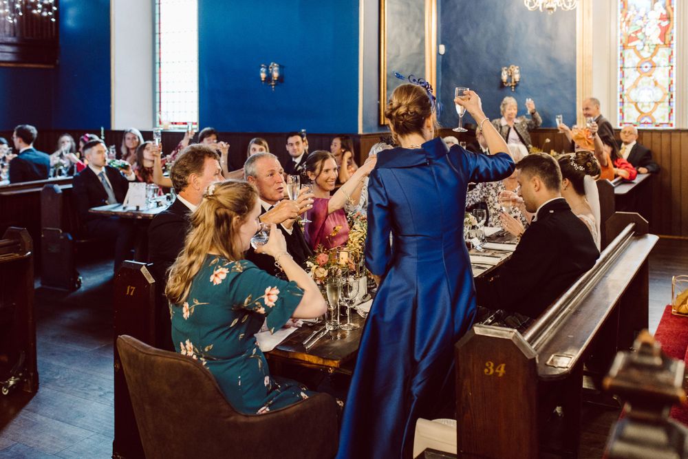 Panoramic view of wedding guests dining in an elegant blue-walled room with stained glass windows.