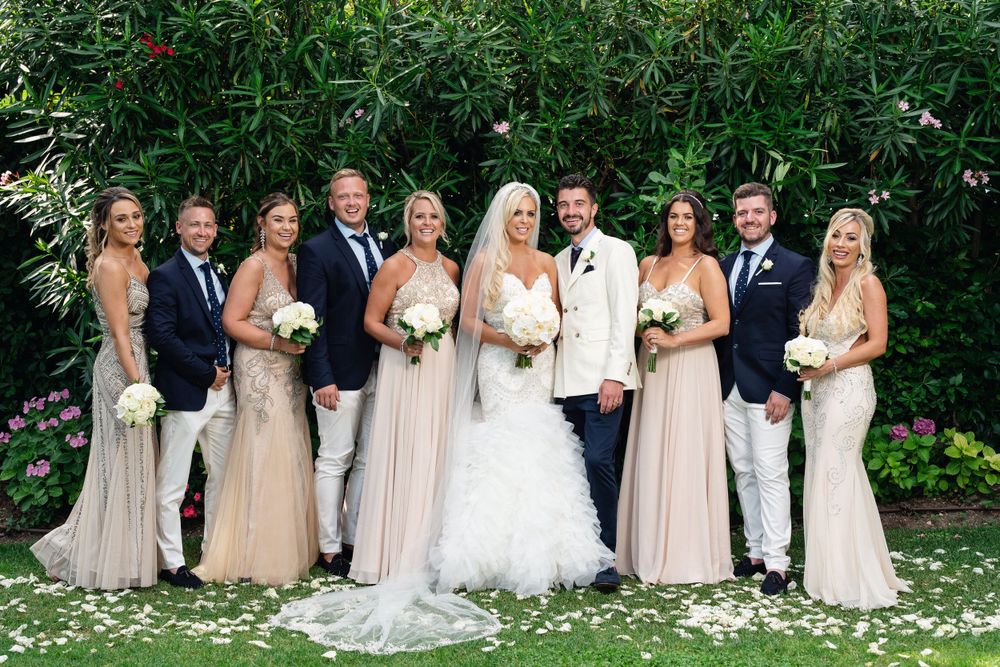 Wedding party poses outdoors in coordinated neutral dresses against a green garden backdrop.