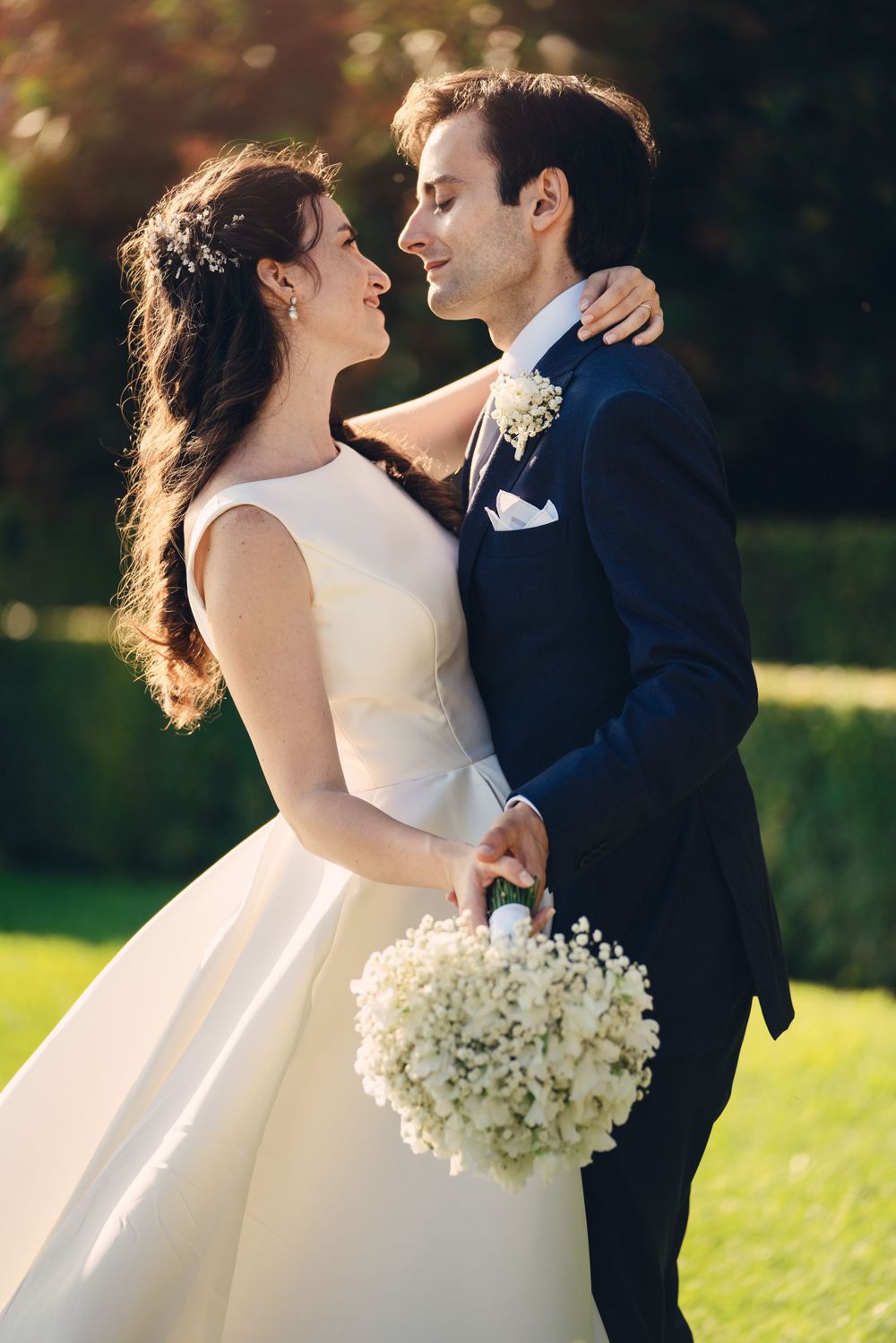 Wedding couple embraces in sunlit garden while holding white floral bouquet.