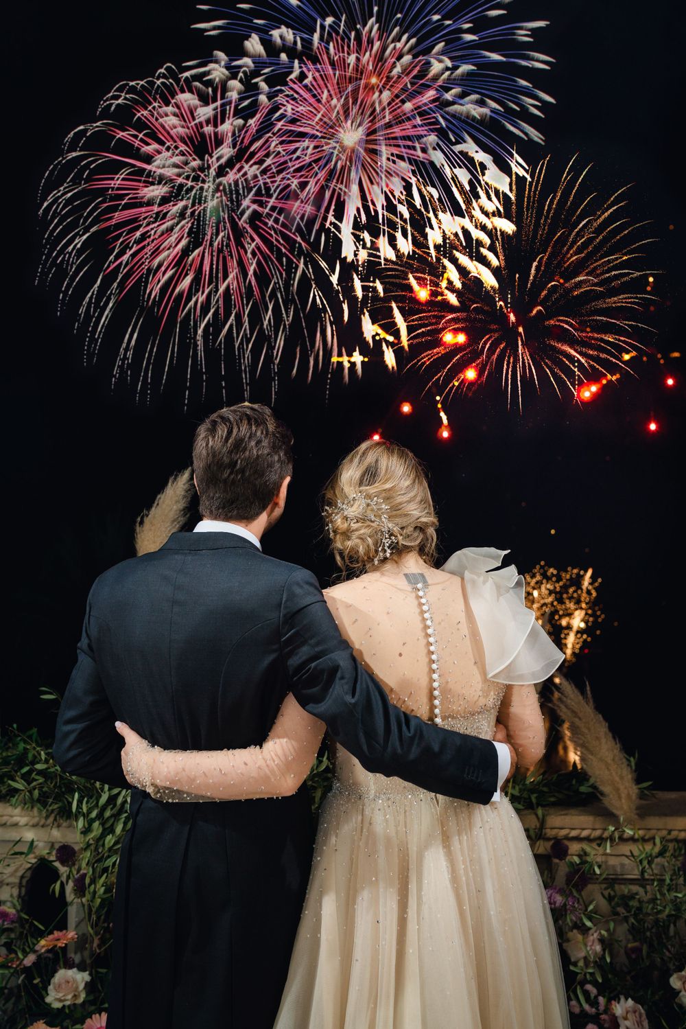 Newlyweds watch fireworks display together during nighttime wedding celebration.