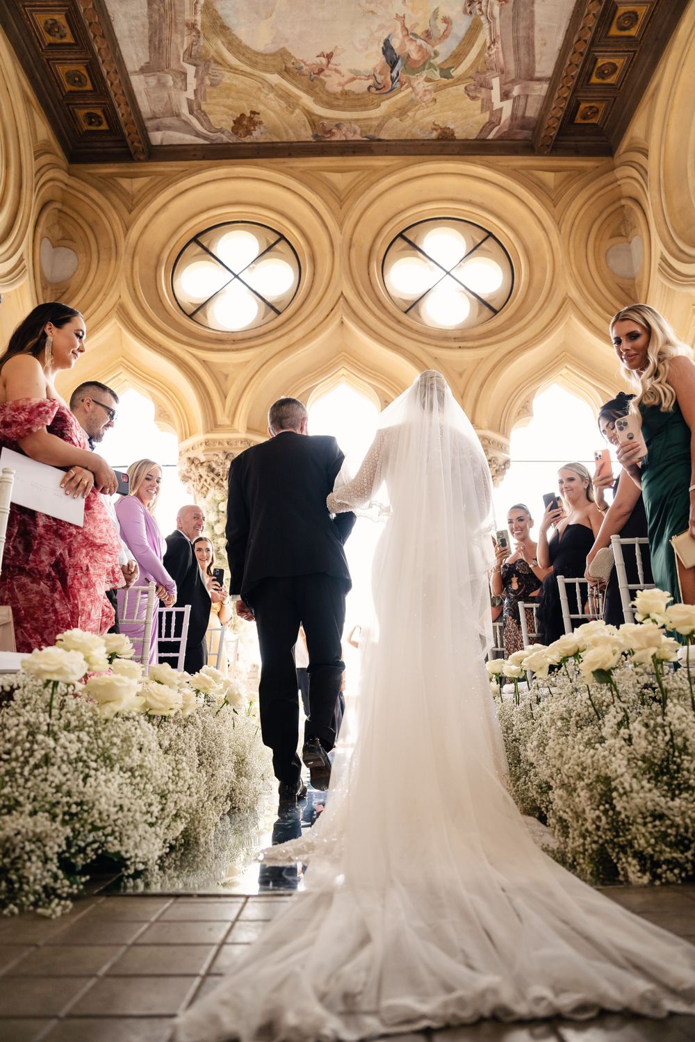 Wedding ceremony in an ornate hall with elegant architecture and white floral decorations lining the aisle.