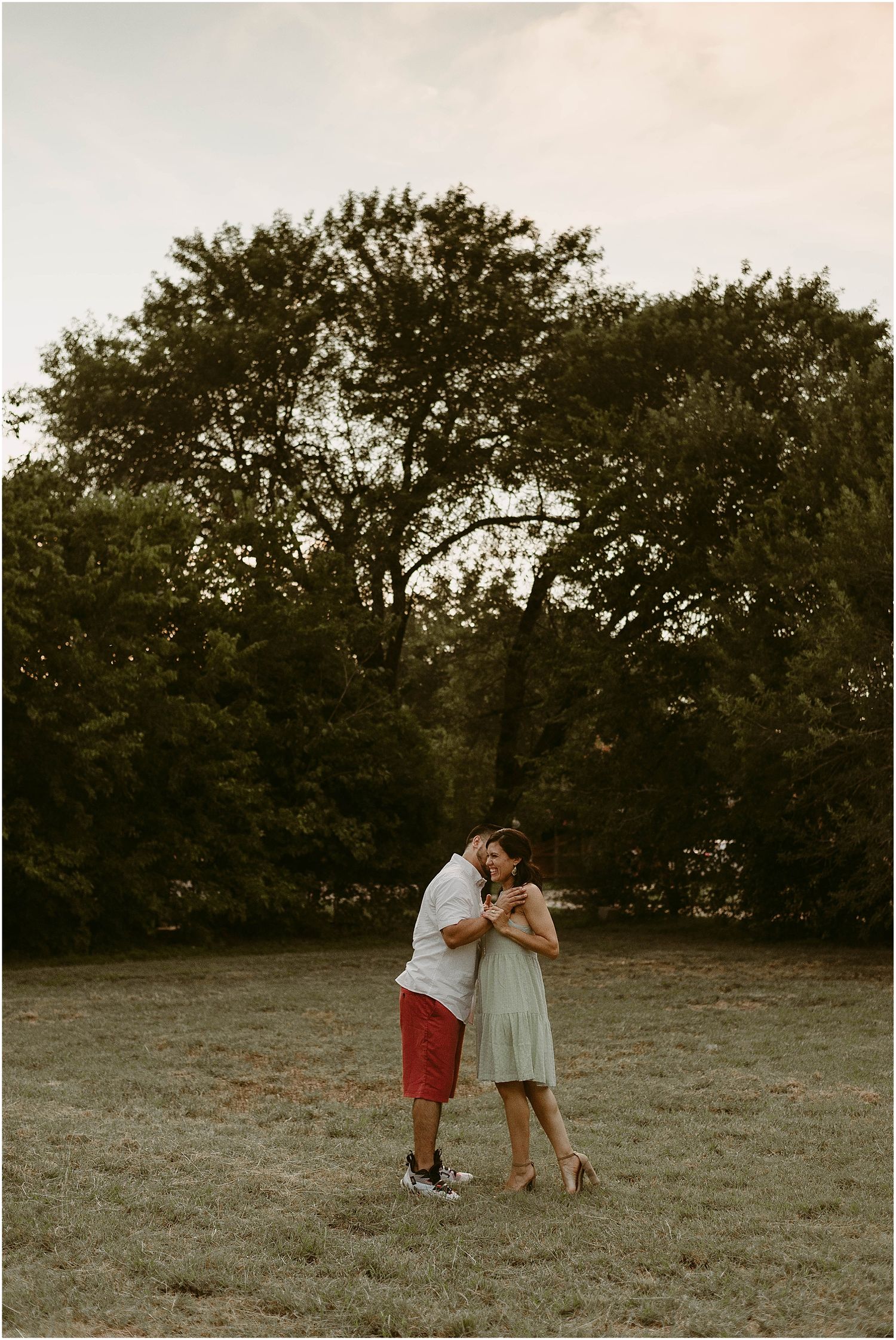 A couple embracing in a field with tall trees silhouetted against the evening sky.