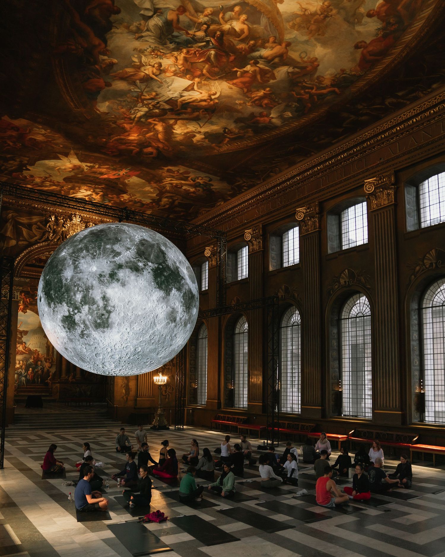 A large moon installation illuminates a grand hall with baroque ceiling frescos as people sit in meditation.
