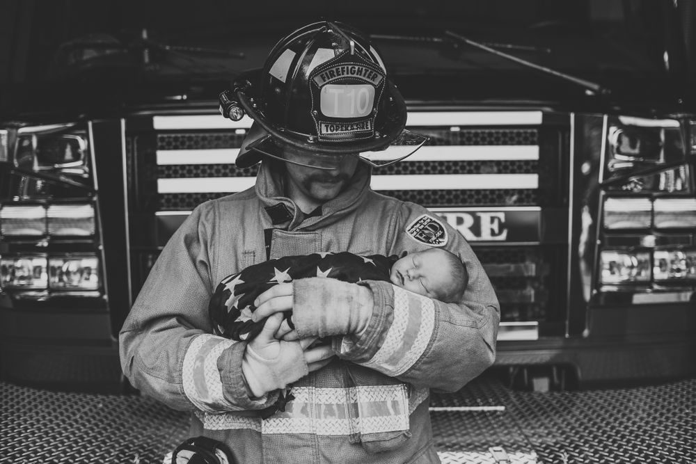 Firefighter in uniform and helmet tenderly holds a newborn baby wrapped in an American flag in black and white.