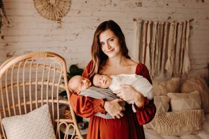 Brunette mother in red dress holding two newborn babies in a boho-styled studio.