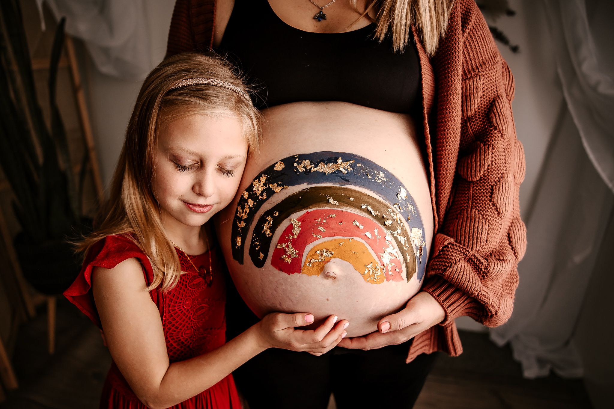 Young girl in red dress touching a painted rainbow baby bump belly art on her mother.