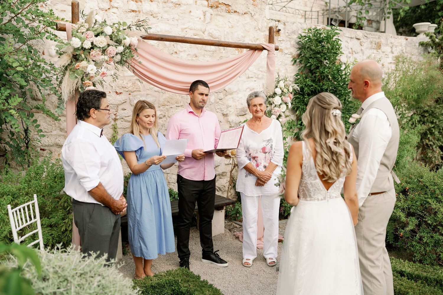Small wedding ceremony gathering under a pink draped arch against a rustic stone wall.