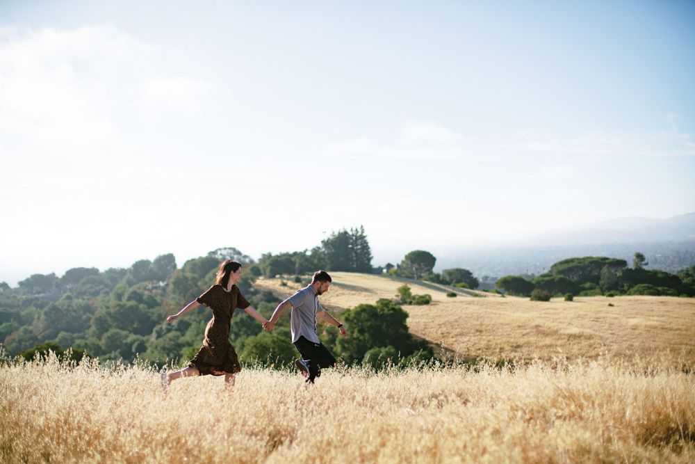 People playfully running through a sun-soaked meadow with rolling hills behind them.