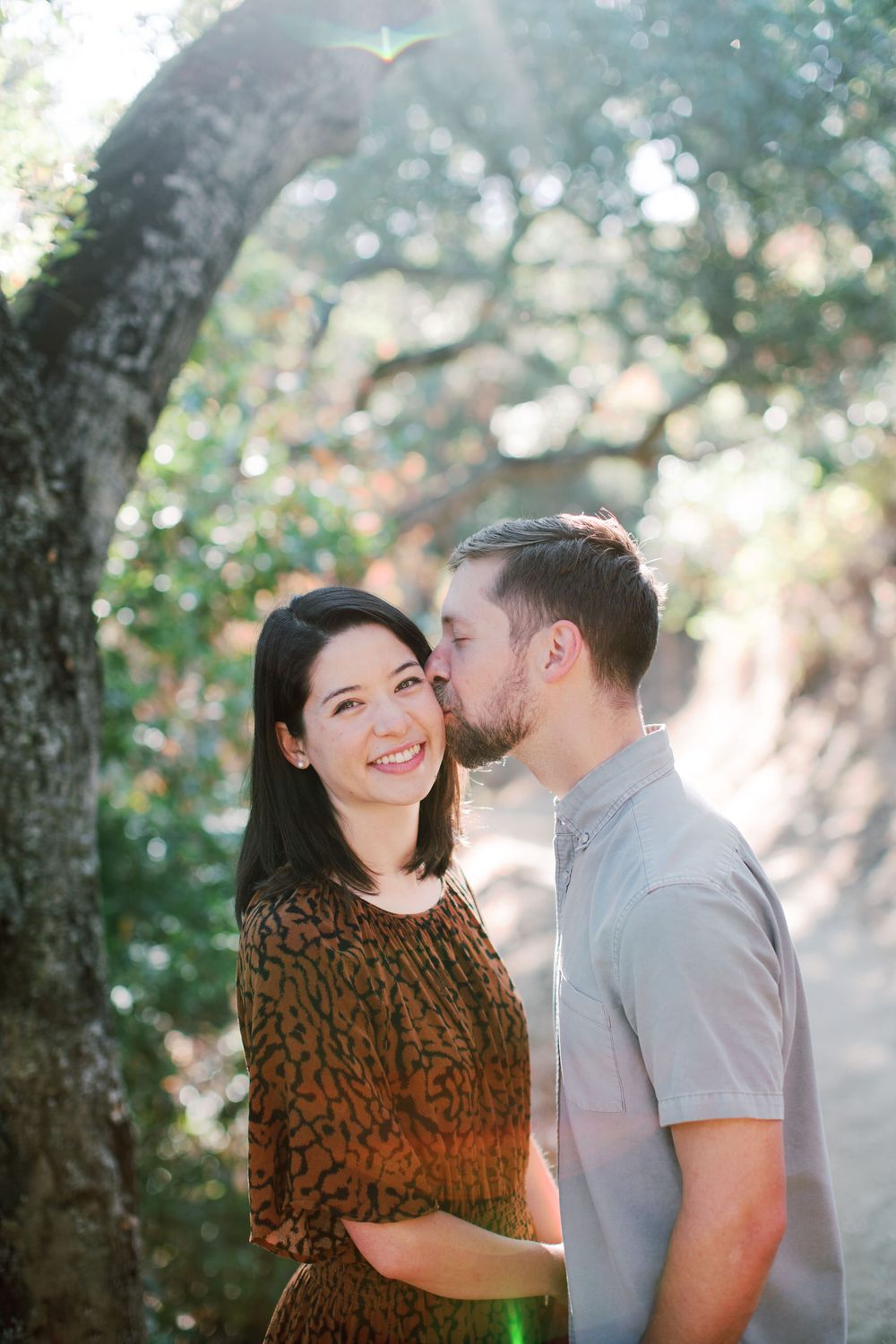 Couple sharing an intimate moment under a large tree with sun flares peeking through.