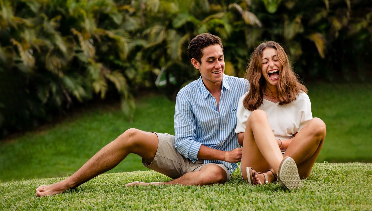 Couple laughing together while sitting on grass in a garden setting.