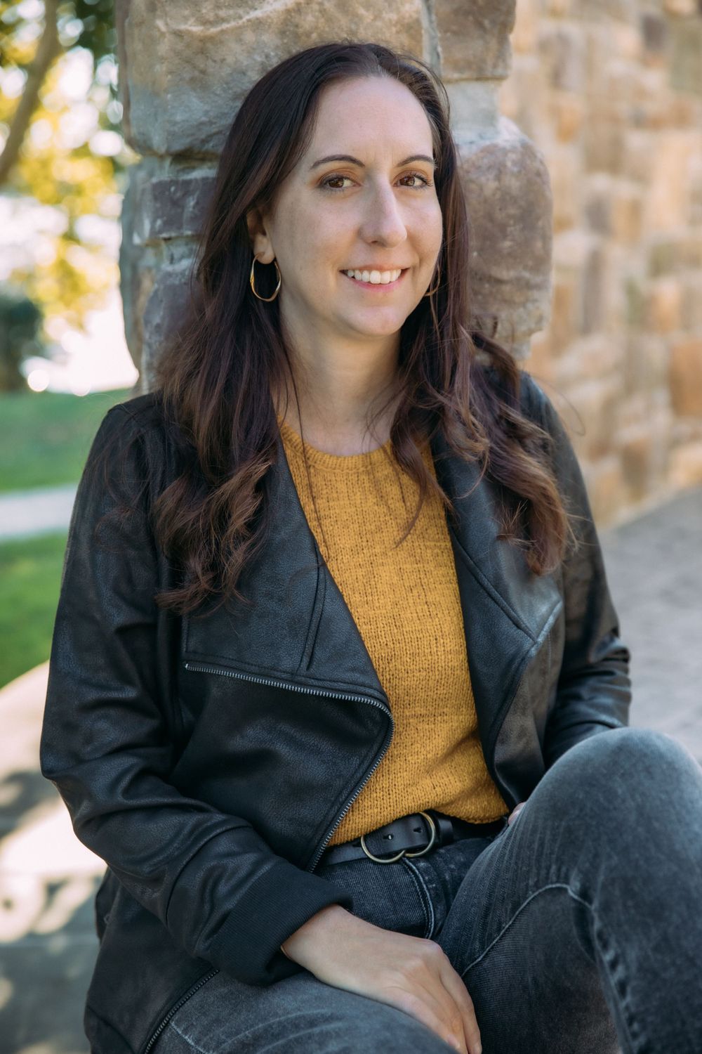 Casual photograph of a woman with brown hair sitting wearing a golden yellow sweater and black leather jacket