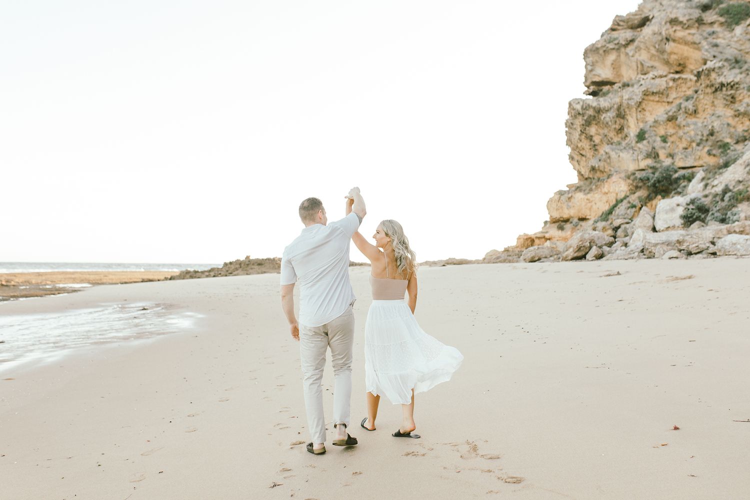 Two people walking hand in hand on a beach with towering cliffs beside them.