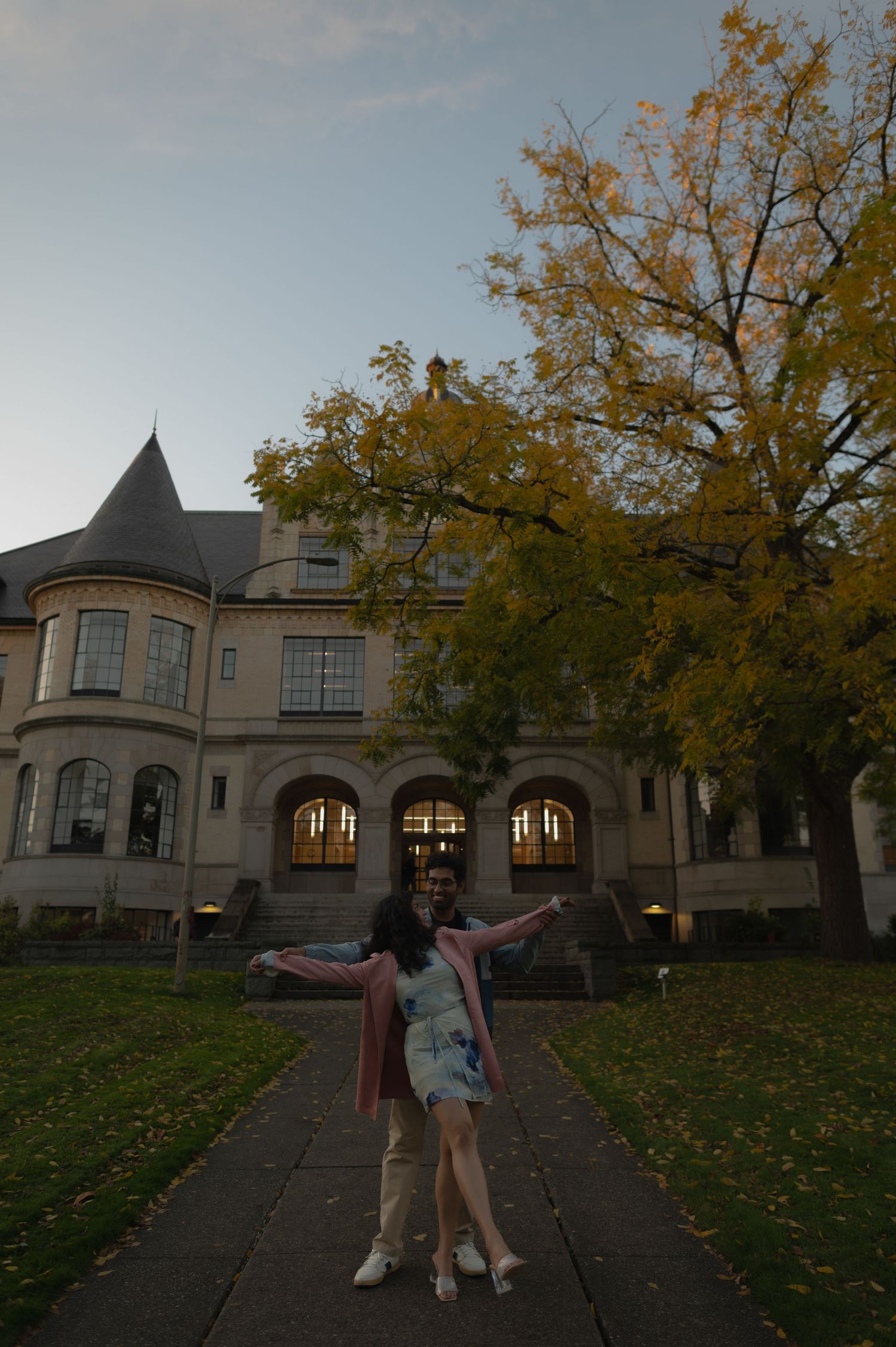 Person poses with arms spread wide in front of a Victorian-style mansion with fall foliage.