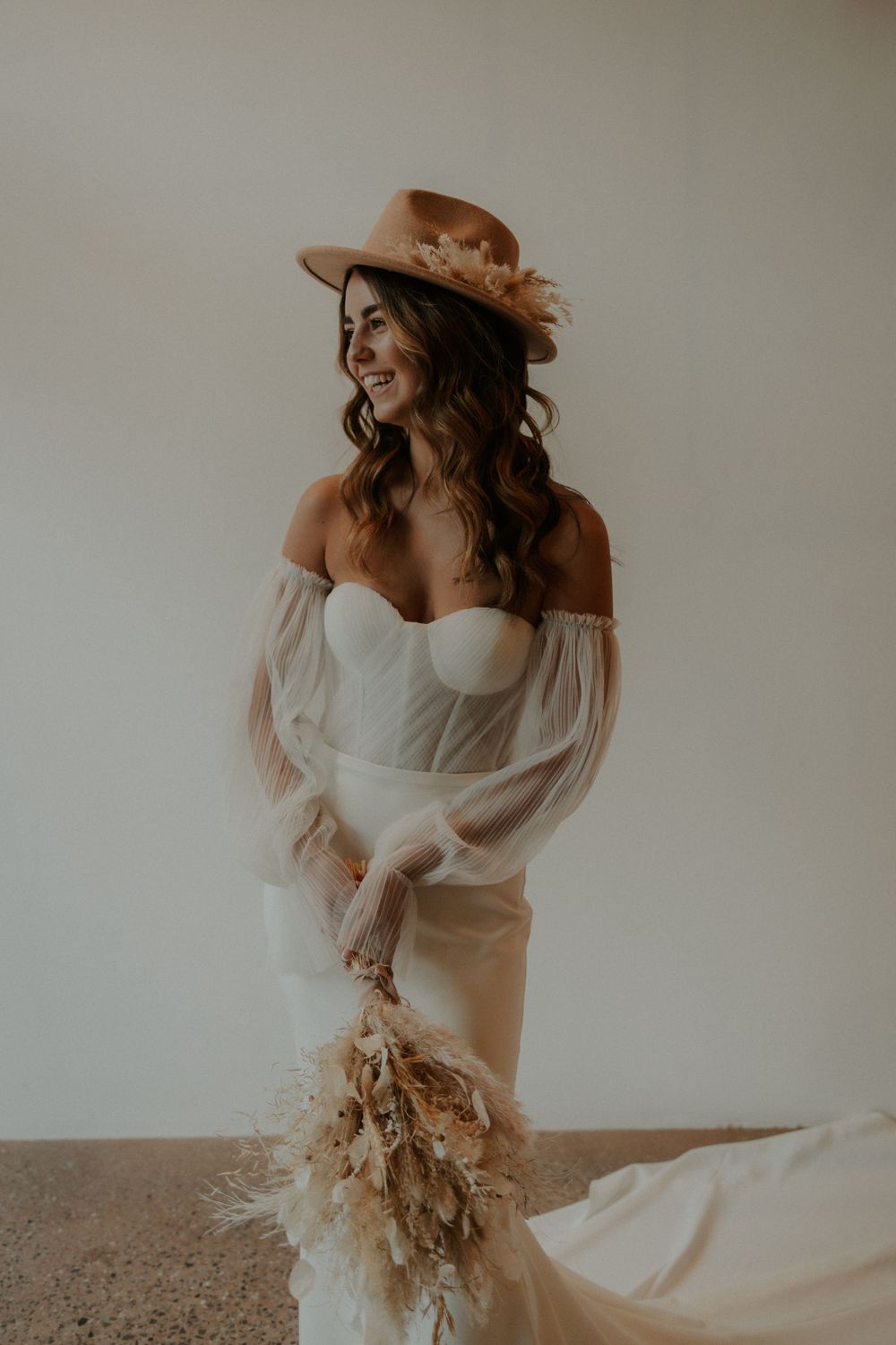 A bride in an off-shoulder dress and brown hat poses with dried flowers.