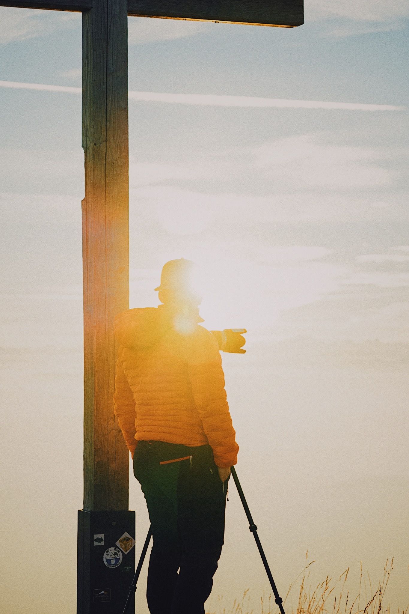 Silhouette of photographer with tripod against golden sunset beside wooden cross.