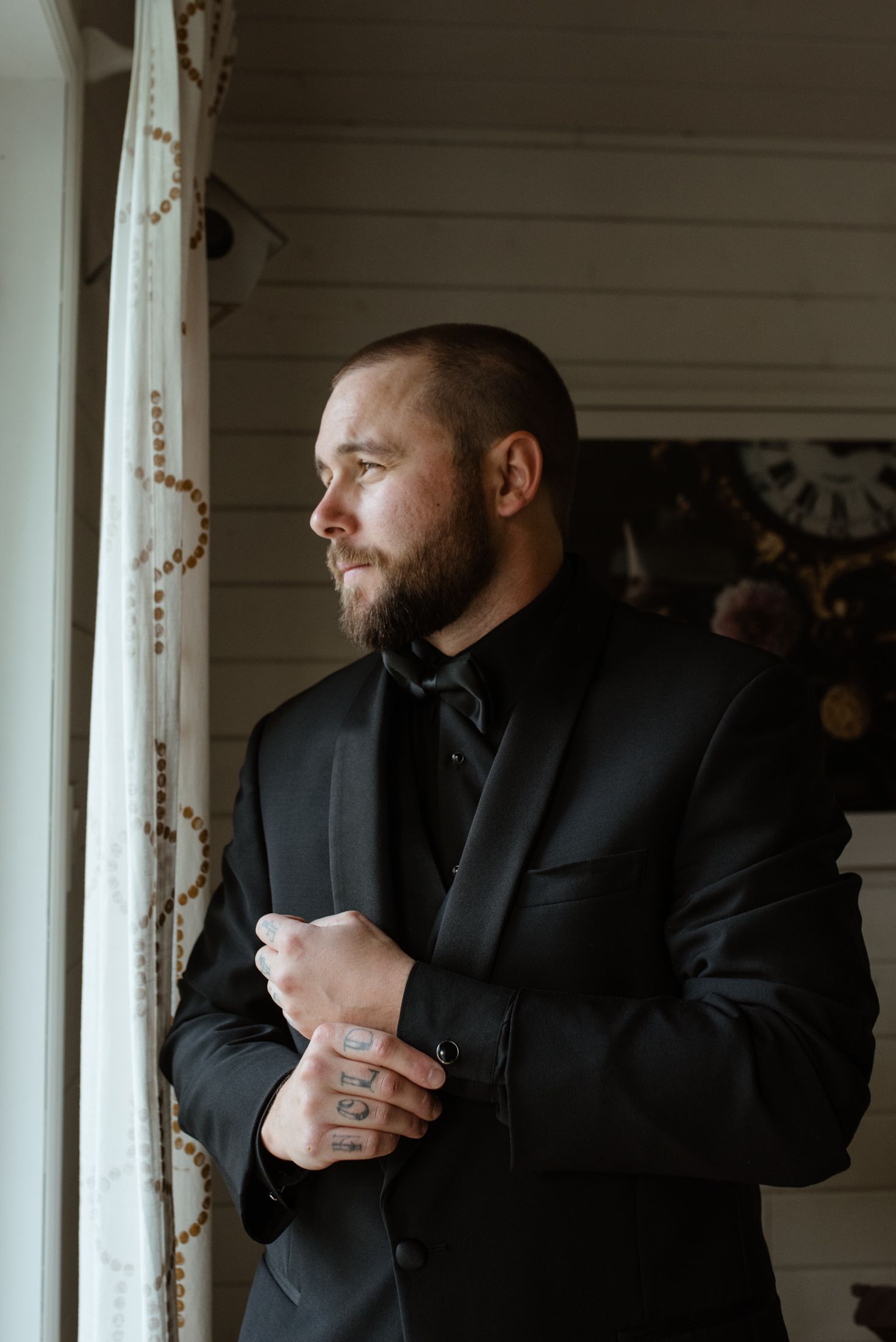 A groom in a black suit adjusts his cuff while standing by a window in moody lighting.