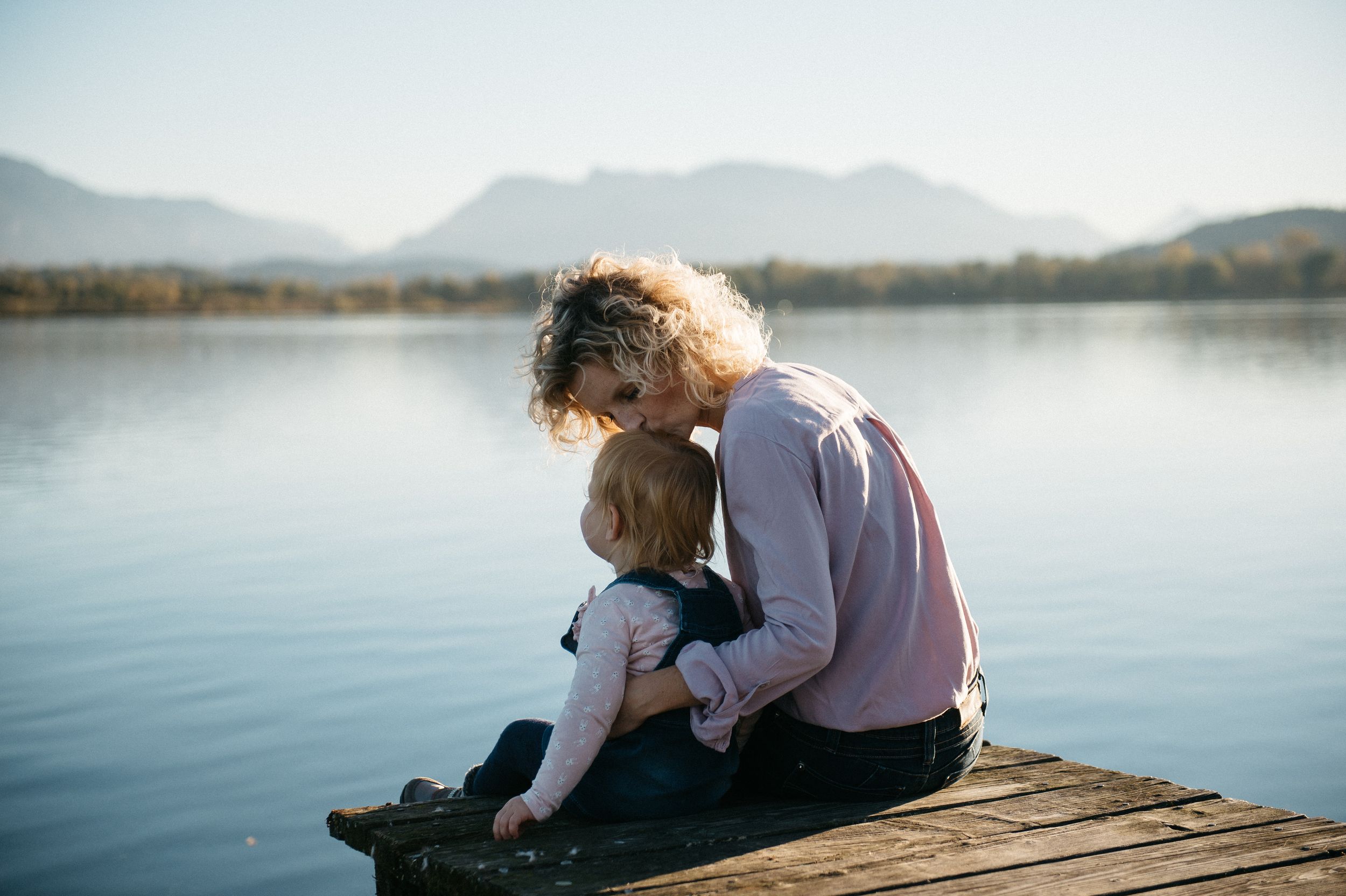 Mutter sitzt mit Tochter innig am Steg und gibt ihr einen Kuss auf den Kopf, im Hintergrund der Chiemsee und Berge