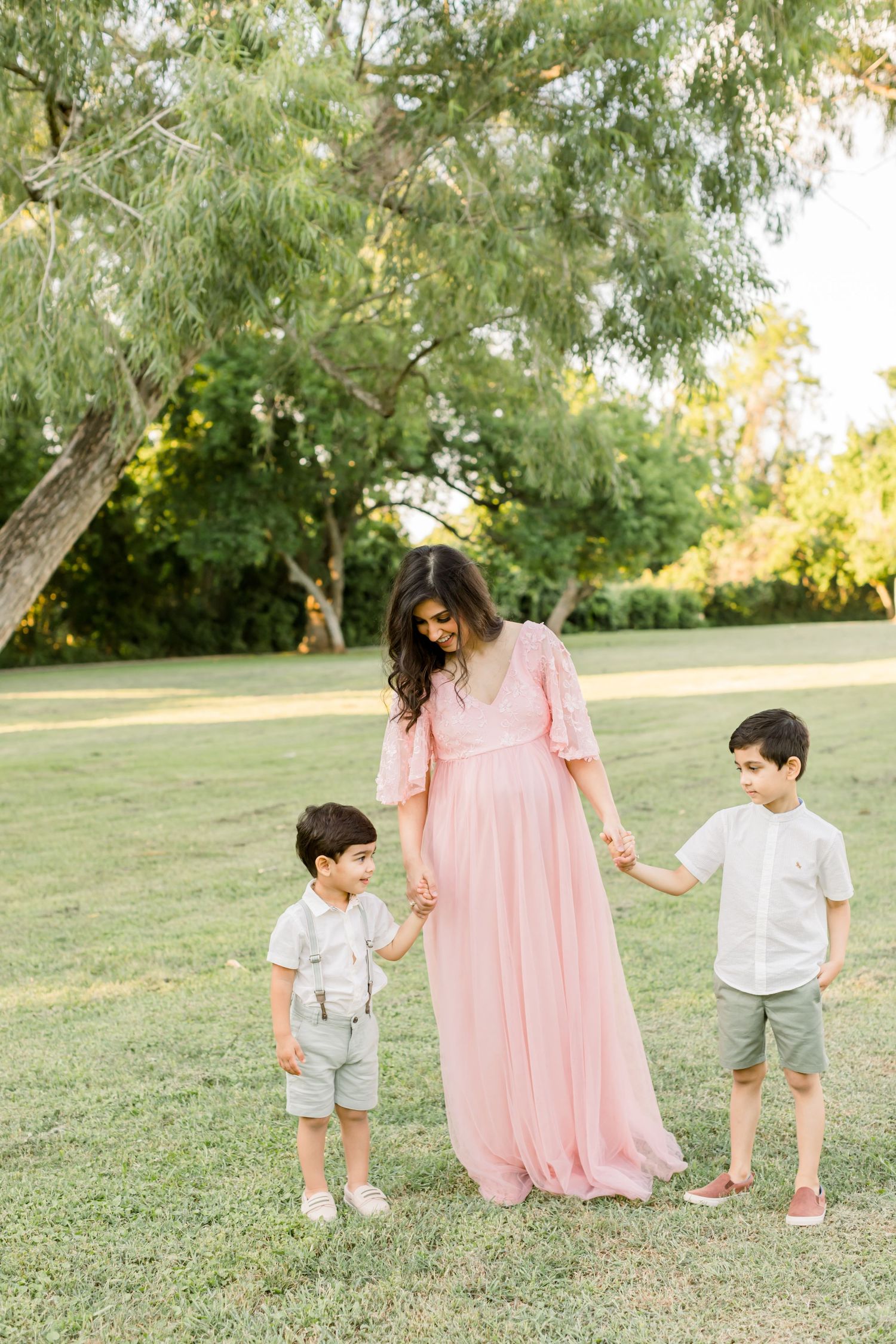 A woman in a pink maxi dress holds hands with two young boys in an outdoor setting.