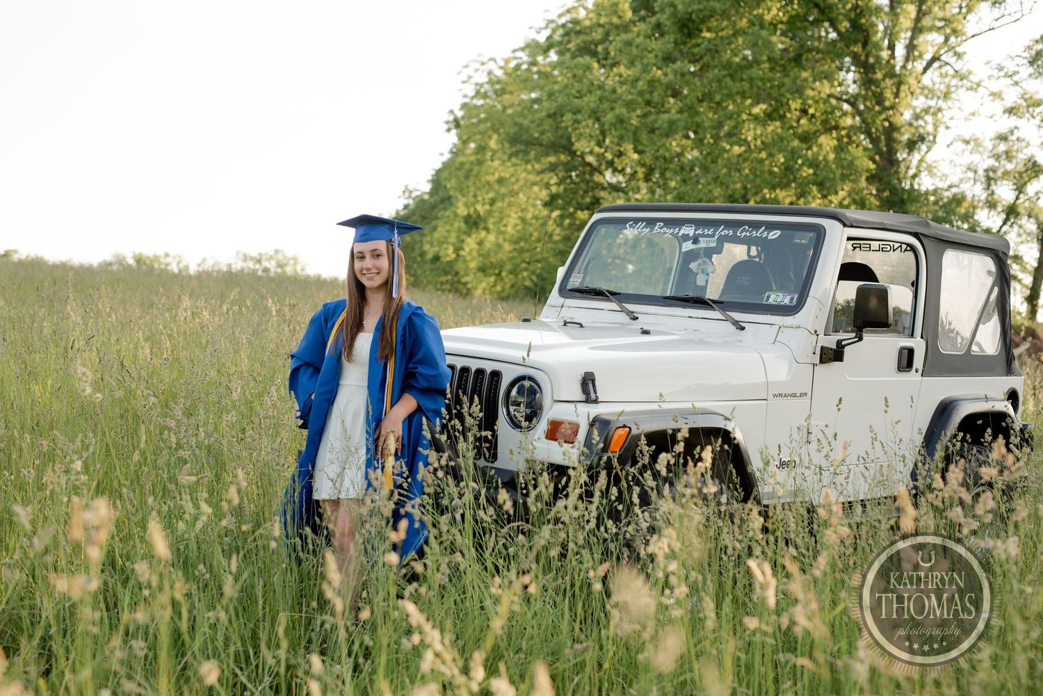 Graduate in blue cap and gown poses in tall grass next to white Jeep.