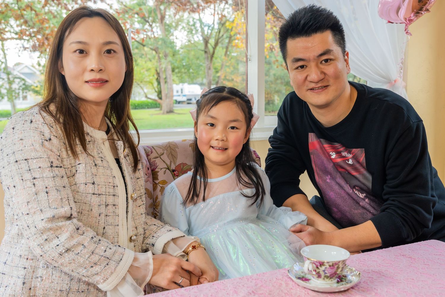 Asian family of three smiling at princess tea party table with floral teacup in cozy room.