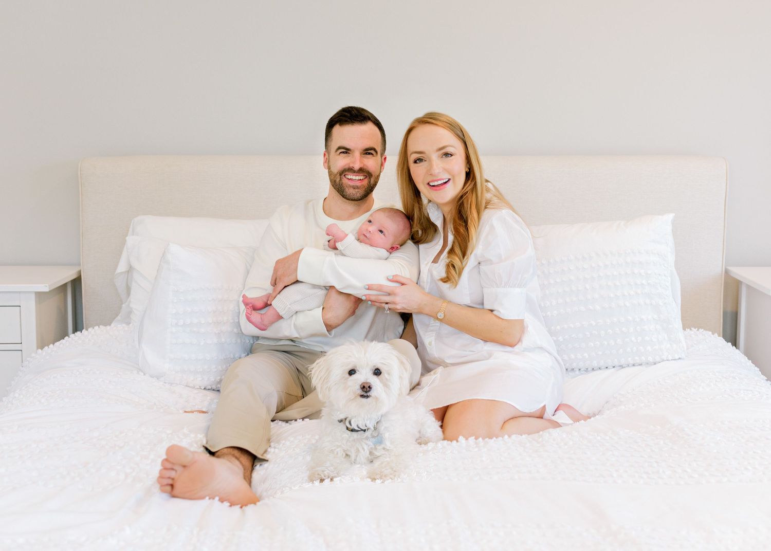 Family poses on white bed with small white dog in bright bedroom setting.