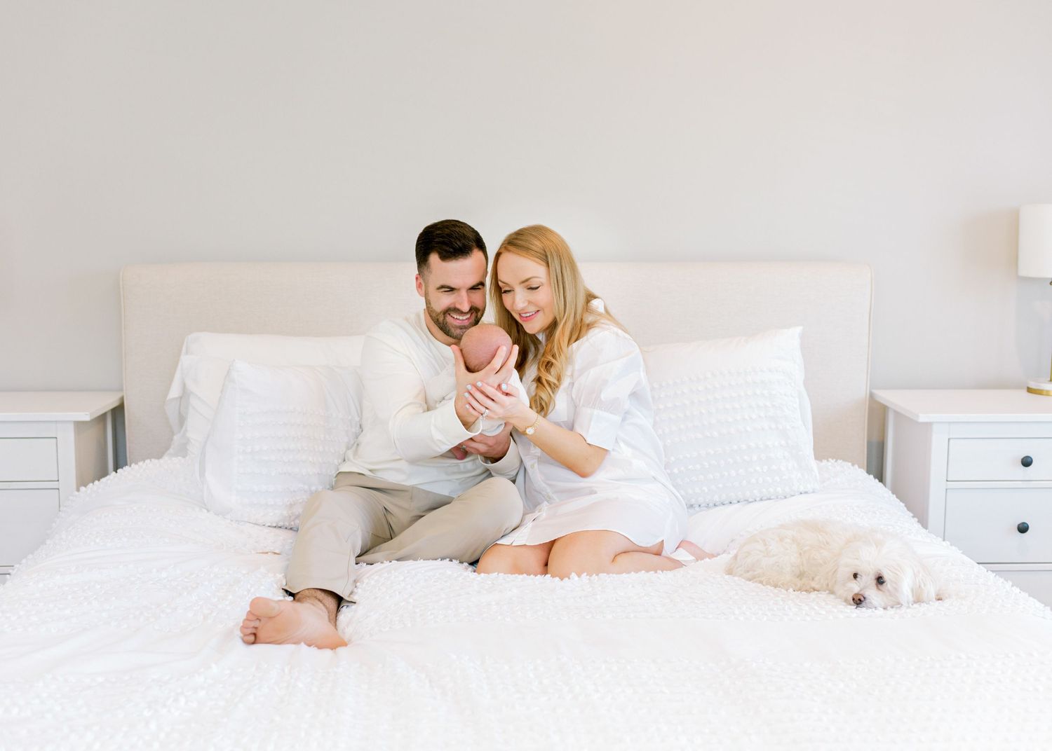 Family sitting together on white bedding with small white dog enjoying tender moment with newborn baby.