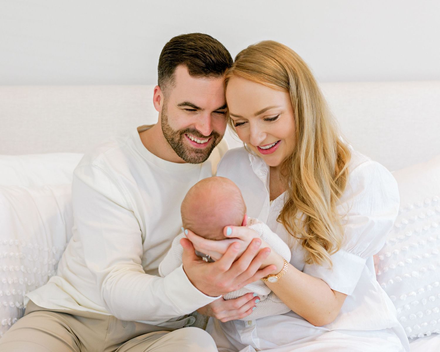 Parents sharing intimate moment with newborn baby on white bed in bright room.